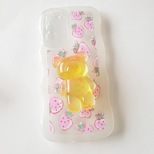 May include: Clear phone case featuring a strawberry pattern and a yellow bear-shaped decoration. The case has a transparent design with pink strawberries and green leaves. The bear is a bright yellow colour.