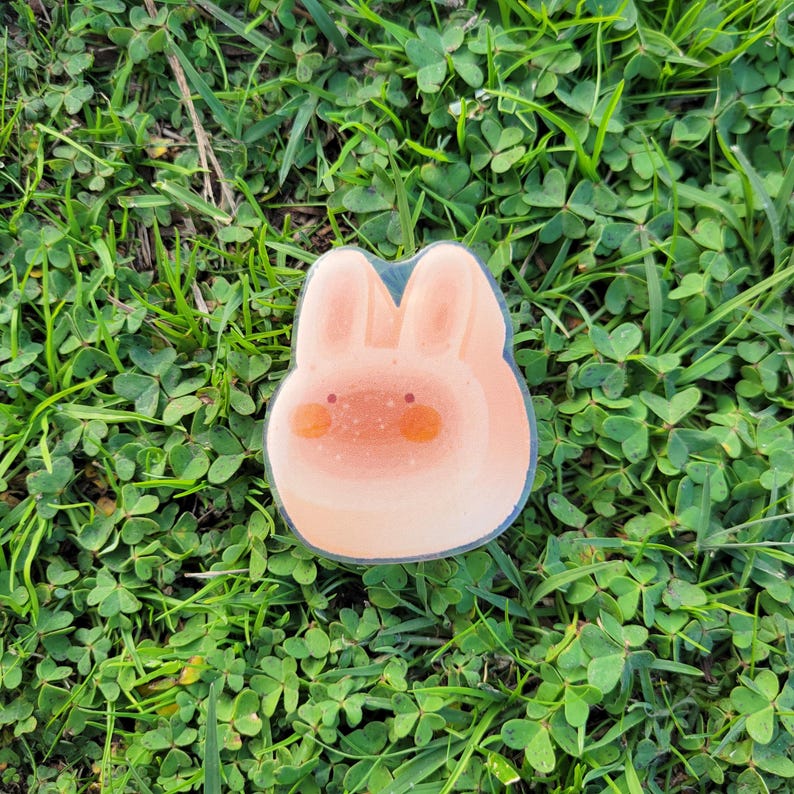 May include: A light pink bunny-shaped sticker with orange cheeks and a sparkly texture. The sticker is outlined in a dark blue color and is set against a background of green grass and clover.