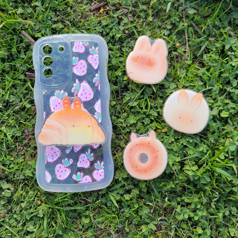 May include: A clear phone case with a strawberry pattern and a croissant-shaped charm. Also, there are three small, round, cartoon-style charms: a bunny, a bunny with orange ears, and a donut.