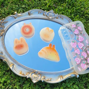 May include: A decorative oval mirror with a silver and gold frame, reflecting four food-shaped resin charms and a clear phone case with a pink strawberry pattern. The charms include a donut, two bunny-shaped treats, and a croissant.