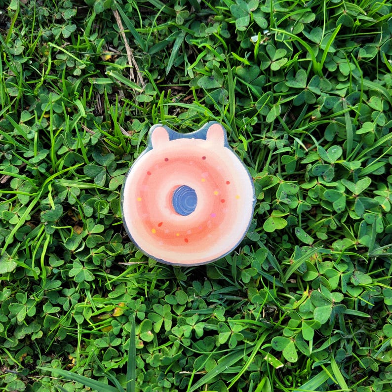 May include: A whimsical donut-shaped pin with a light pink frosting and colorful sprinkles. The pin has a blue center and a small, rounded top, resembling ears. The pin is set against a backdrop of green grass and clover.