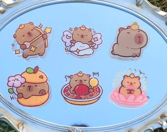 May include: Six cartoon capybara stickers in different poses. One has a bow and arrow, another angel wings, and others are in fruit, a pizza, and a lotus flower. The stickers are displayed on a reflective surface within a decorative frame.