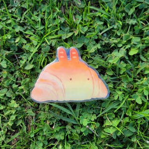 May include: A whimsical sticker featuring a cartoon croissant with orange and yellow gradients. The croissant has two orange ears and two small brown dots for eyes. The sticker is set against a backdrop of green grass and clover.