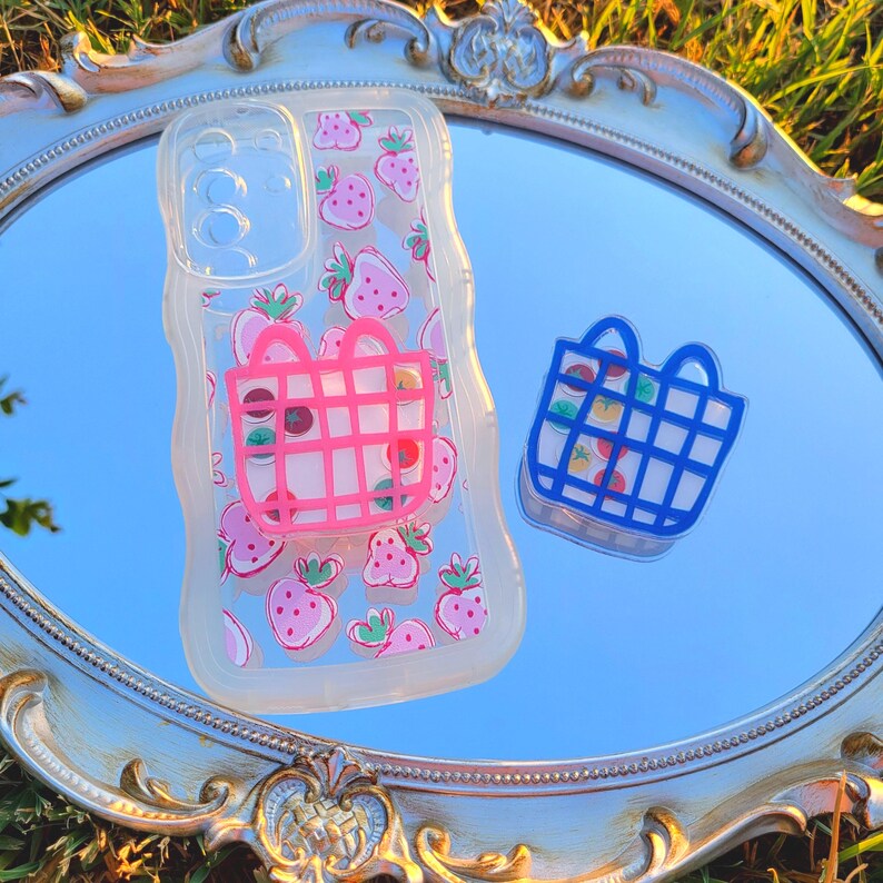 May include: Clear phone case with a strawberry pattern and a pink shopping basket design. A blue shopping basket sticker is next to the case. The items are on a decorative mirror.