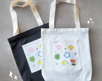 Puede incluir: Dos bolsas de lona con un diseño floral. Una bolsa es negra con un bolsillo blanco y la otra es blanca con un bolsillo negro. Ambas bolsas tienen un diseño floral colorido con flores, hojas y una pequeña flor roja con una cara.