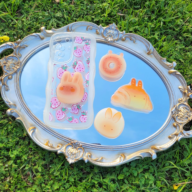 May include: A clear phone case with a strawberry pattern and a 3D bunny. Also, a donut, croissant, and bunny-shaped charms. The items are arranged on a decorative silver-framed mirror.