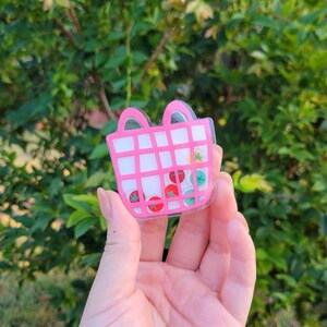 May include: A pink and white shopping basket-shaped charm with a grid design. The charm contains small, colourful, fruit-shaped confetti pieces. The charm is held against a green leafy background.