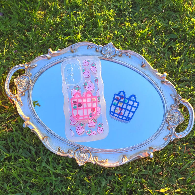 May include: A clear phone case featuring a pink strawberry pattern and a pink basket design. A small blue basket-shaped sticker is next to the phone case. The items are on a silver-framed mirror, set against green grass.