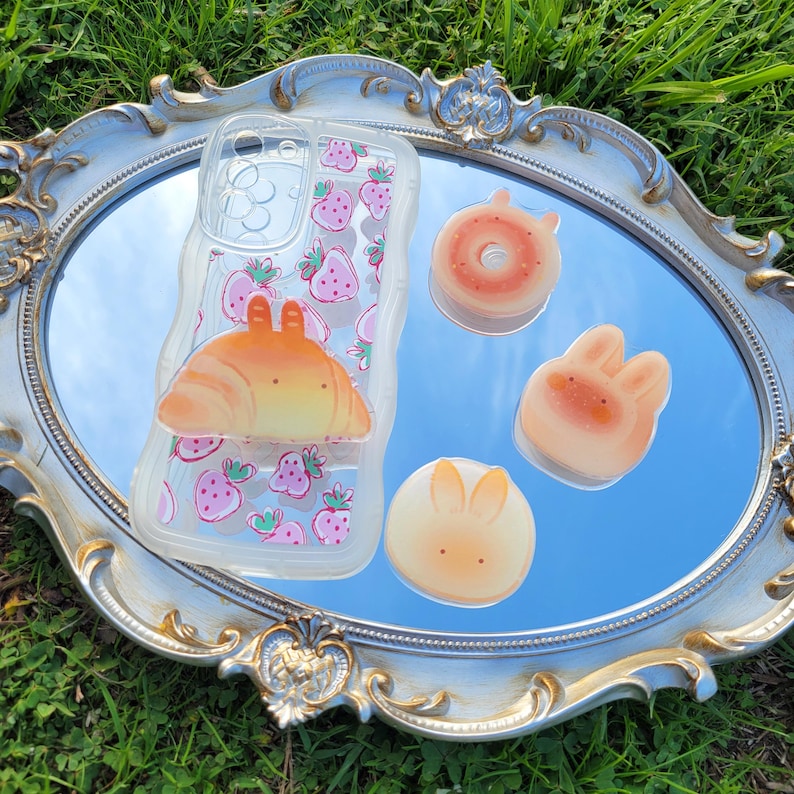 May include: A clear phone case with a pink strawberry pattern and a croissant charm. Beside the phone case are three food-shaped charms: a donut, and two bunny-shaped treats. The items are arranged on a silver-framed mirror.