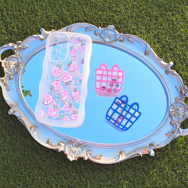 May include: A clear phone case with a pink strawberry pattern, a pink basket with colourful beads, and a blue basket with colourful beads are displayed on a light blue oval mirror. The mirror is on a green surface.