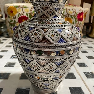May include: A silver and ceramic vase with a geometric pattern in shades of blue, green, brown, and white. The vase has a wide base and a narrow neck.