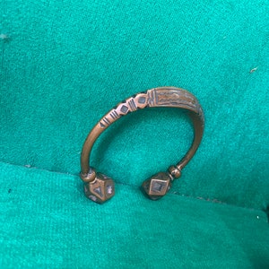 May include: A bronze bracelet with a geometric design and two decorative ends. The bracelet is on a green surface.