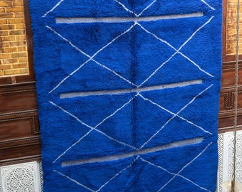 Puede incluir: Una alfombra azul con un patrón de diamantes blancos. La alfombra tiene un borde de flecos y está colgada en una pared.