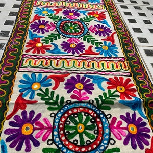 Peut inclure: Chemin de table brodé avec un fond crème et des motifs floraux colorés. Le design présente de grandes fleurs rouges, bleues, violettes et roses, avec des feuilles vertes et une bordure décorative. Mesure environ 180 cm de long.
