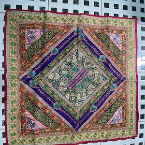 May include: A square tapestry with a diamond-shaped pattern in the center. The tapestry is made of different fabrics and colors, including red, green, blue, and gold. The edges of the tapestry are trimmed with gold thread.
