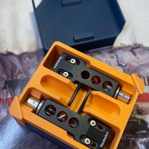 Könnte beinhalten: Eine orangefarbene und marineblaue Aufbewahrungsbox enthält zwei schwarze Technics-Tonabnehmer. Die Tonabnehmer haben silberne Anschlüsse und rote und schwarze Kabel. Die Box ist offen, mit dem Deckel dahinter.