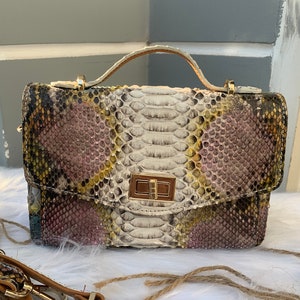 Pode incluir: Uma pequena bolsa branca com um padrão de pele de cobra em tons de rosa, amarelo e verde. A bolsa tem uma alça superior dourada e um fecho de encaixe dourado. Acompanha uma alça de couro marrom com um fecho dourado.