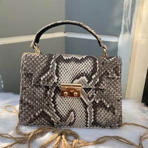 Puede incluir: Un pequeño bolso de mano de piel de serpiente marrón y blanco con cierre dorado y asa negra. El bolso está sobre una superficie blanca y esponjosa con una cuerda marrón atada en un lazo.