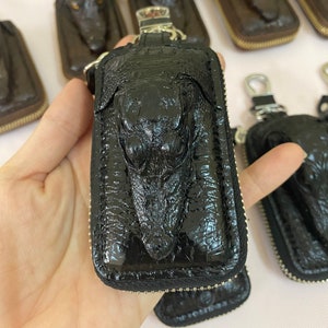 Könnte beinhalten: Schwarzer Leder-Schlüsselanhänger mit Krokodilkopf-Design. Der Schlüsselanhänger hat einen Reißverschluss und einen Metallring.