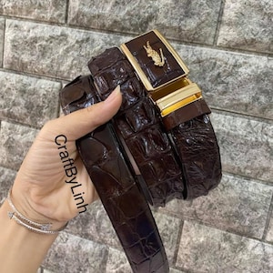 May include: A dark brown leather belt with a crocodile skin texture. The rectangular buckle has a gold-coloured frame and a gold-coloured animal emblem. The text "CraftByLinh" is visible on the left side of the image.