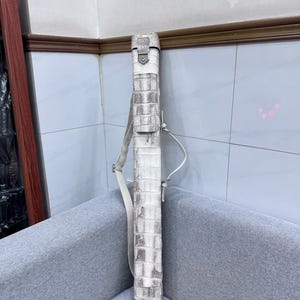May include: A tall, white, rectangular bag with a textured, crocodile-like pattern. It has a shoulder strap and a buckle closure at the top. The bag is standing upright on a gray surface.