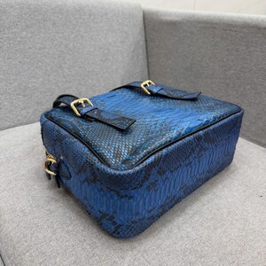 May include: A rectangular crossbody bag featuring a vibrant blue and black snakeskin pattern. The bag includes two top straps with gold-toned metal buckles and a gold-toned metal side ring attachment, showcasing a structured design with black piping.