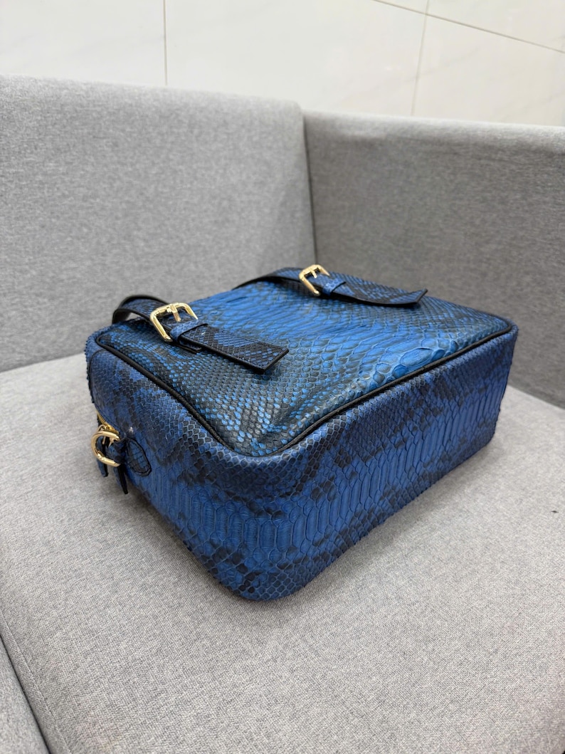 May include: A rectangular crossbody bag featuring a vibrant blue and black snakeskin pattern. The bag includes two top straps with gold-toned metal buckles and a gold-toned metal side ring attachment, showcasing a structured design with black piping.