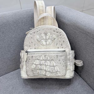 Könnte beinhalten: Ein weißer Rucksack mit einer strukturierten, schuppenartigen Oberfläche. Der Rucksack verfügt über einen Tragegriff, zwei verstellbare Schultergurte, ein Hauptfach mit Reißverschluss sowie eine kleinere Fronttasche mit silbernen Beschlägen.