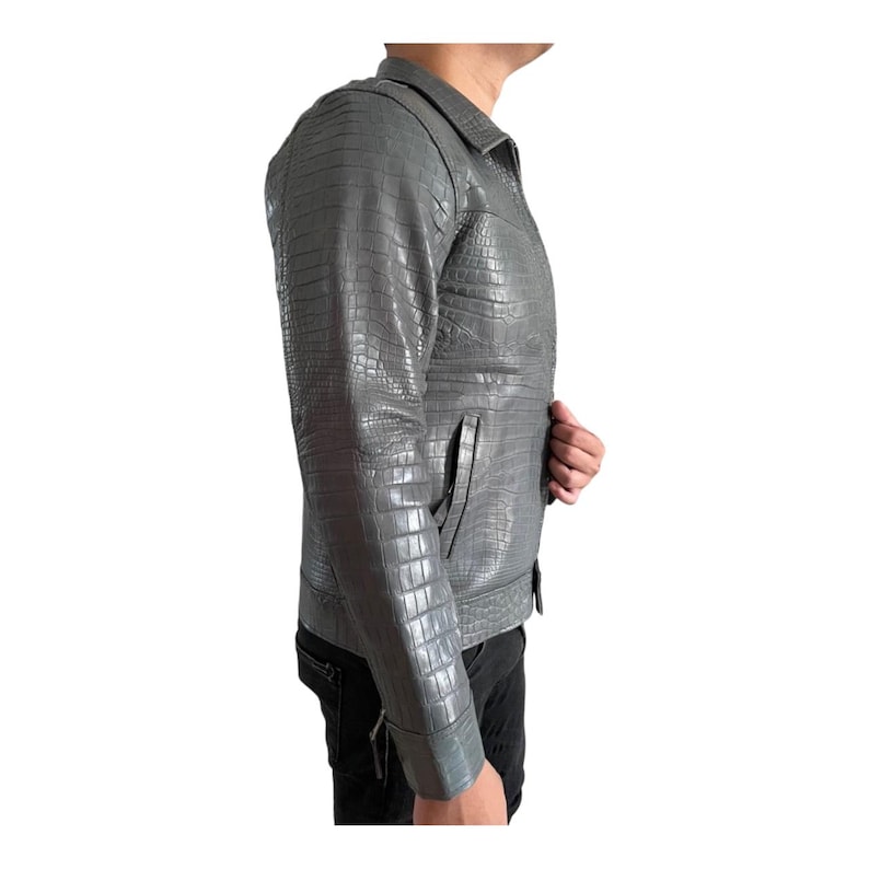 May include: A gray, textured leather jacket with a crocodile pattern. The jacket has a collared neckline, a front pocket, and a zipper closure. The jacket is worn by a person in black pants.