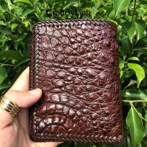May include: A brown leather wallet with a crocodile skin pattern and a braided edge. The wallet is closed and has a stitched design.
