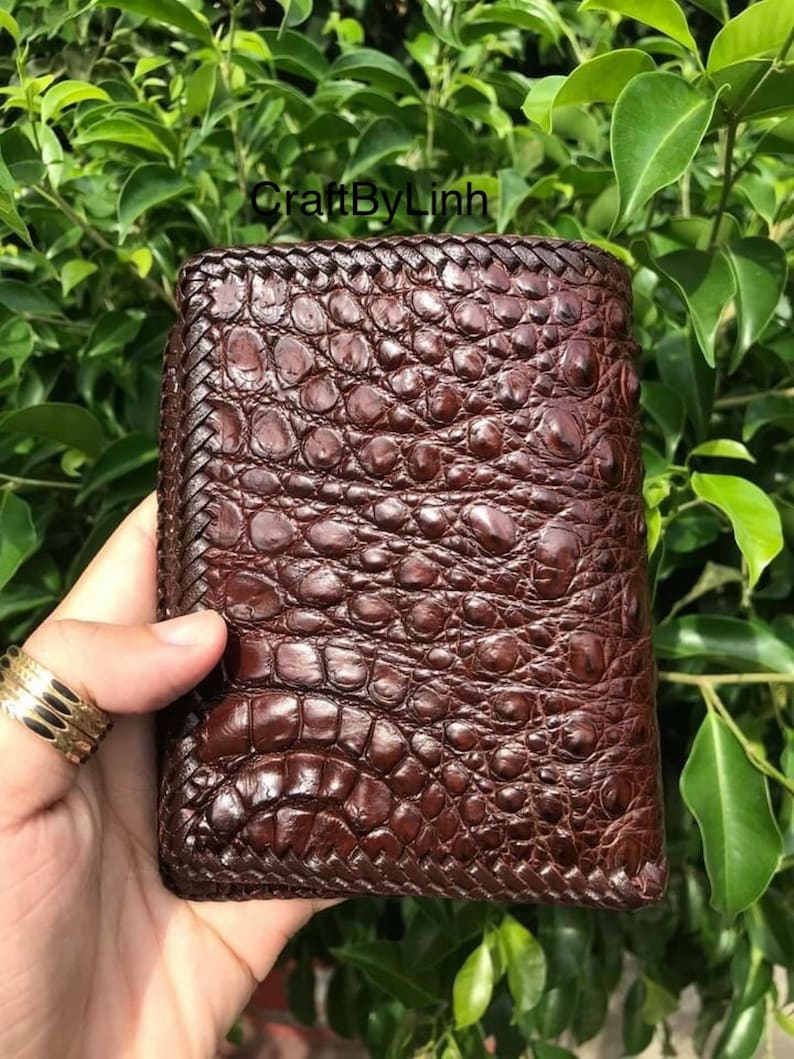 May include: A brown leather wallet with a crocodile skin pattern and a braided edge. The wallet is closed and has a stitched design.