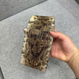 Op de afbeelding: Een bi-fold portemonnee met een gestructureerd, reliëfpatroon dat het uiterlijk van reptielenhuid nabootst. Het oppervlak vertoont een verloop van donkerbruine en crèmetinten, met een opvallend centraal schubbenontwerp dat verticaal over de buitenkant loopt.