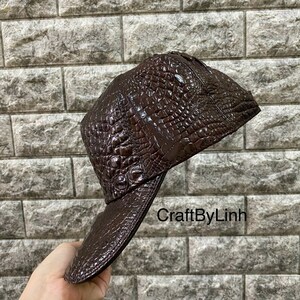 May include: A brown leather baseball cap with a crocodile skin pattern. The cap has a curved brim and a snap closure. The text "CraftByLinh" is visible on the brim.