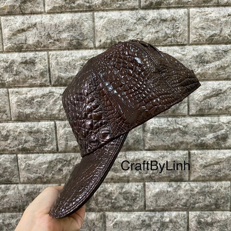 May include: A brown leather baseball cap with a crocodile skin pattern. The cap has a curved brim and a snap closure. The text "CraftByLinh" is visible on the brim.