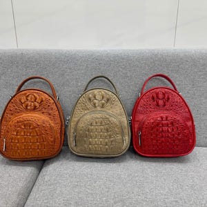 Op de afbeelding: Drie koepelvormige rugzakken met een gestructureerd krokodillenleerpatroon. De rugzakken zijn oranje, metallic goud en rood. Elke tas is voorzien van een handvat aan de bovenkant, een voorvak met rits en een hoofdvak met ritssluiting.