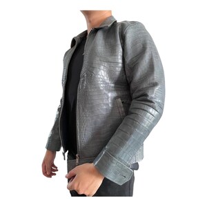 May include: A gray, textured leather jacket with a crocodile pattern. The jacket has a zipper closure, a collar, and a pocket on the chest. The jacket is a neutral color and appears to be made of high-quality leather.