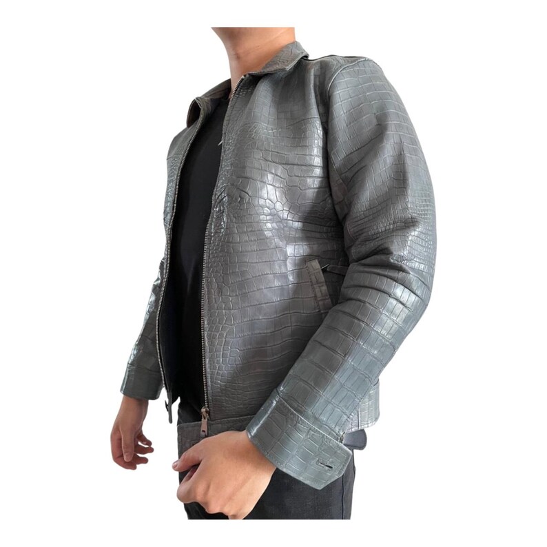 May include: A gray, textured leather jacket with a crocodile pattern. The jacket has a zipper closure, a collar, and a pocket on the chest. The jacket is a neutral color and appears to be made of high-quality leather.