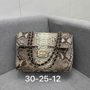 May include: A rectangular handbag featuring a snakeskin pattern in shades of beige, gray, and brown. The bag has a gold-tone metal turn-lock closure and a double chain strap interwoven with black leather. The text 30-25-12 is printed below the bag.