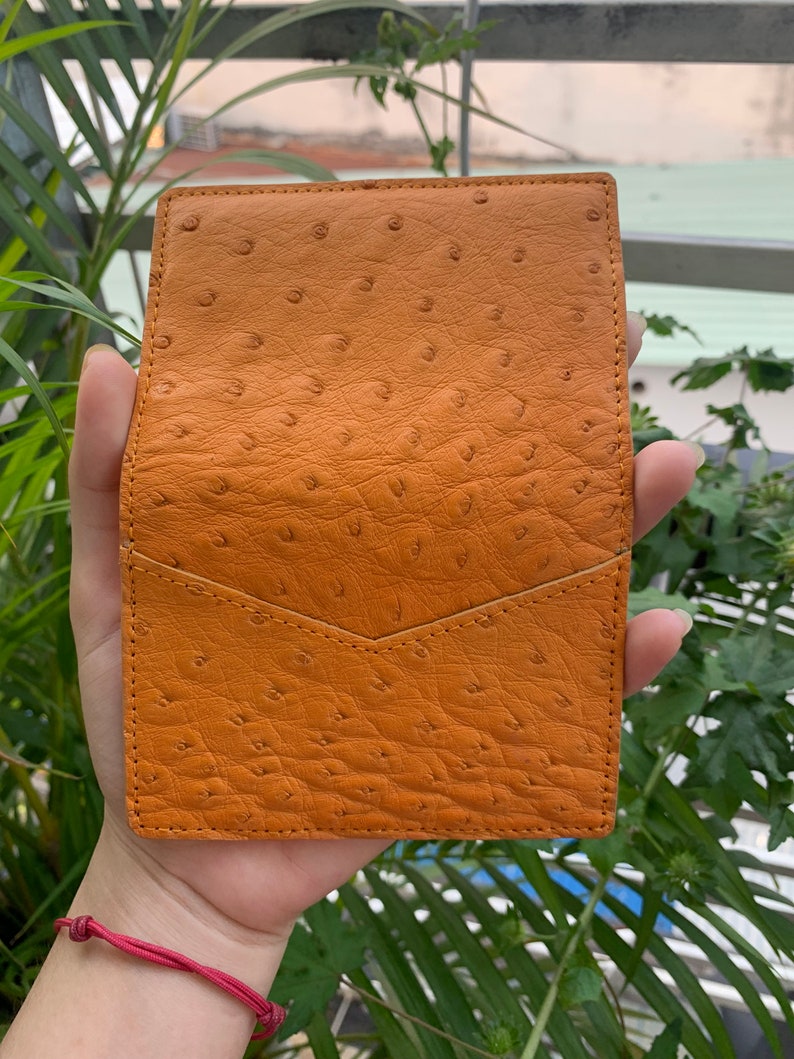 Puede incluir: Una peque&ntilde;a cartera de cuero naranja con un patr&oacute;n texturizado de piel de avestruz. La cartera tiene un cierre de solapa y un solo bolsillo.
