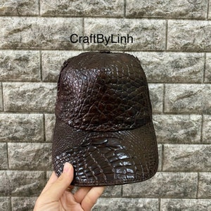 May include: A brown leather baseball cap with a crocodile skin pattern. The cap has a curved brim and an adjustable strap in the back.