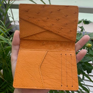 Puede incluir: Una cartera de cuero marr&oacute;n con m&uacute;ltiples ranuras para tarjetas y un cierre de solapa. La cartera tiene un patr&oacute;n texturizado de piel de avestruz.