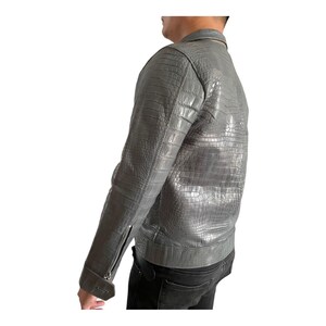 May include: A gray leather jacket with a crocodile skin texture. The jacket has a zipper closure and a collar. The sleeves have zippers at the cuffs. The person is wearing black jeans.