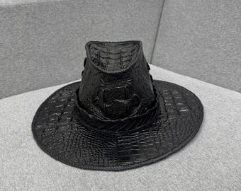 May include: A black fedora-style hat with a wide brim. The hat is made of a textured material, possibly leather, with a crocodile or alligator skin pattern. The hat is resting on a gray surface.