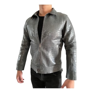 May include: A gray leather jacket with a crocodile skin texture. The jacket has a zipper closure, a collar, and long sleeves. The jacket is worn over a black shirt and black pants.