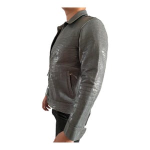 May include: A gray leather jacket with a textured, crocodile-like pattern. The jacket has a collared neckline, a zipper closure, and a front pocket. The sleeves have buttoned cuffs. The jacket is shown from the side.