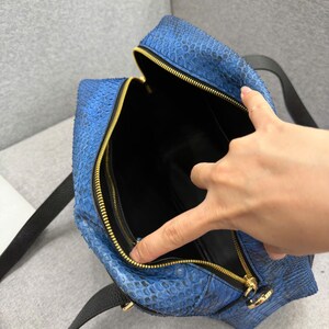 May include: A blue snakeskin-textured handbag featuring a gold-toned zipper closure and black leather straps. The interior of the bag is lined with black material, and the top is open to reveal the main compartment and a zippered side pocket.