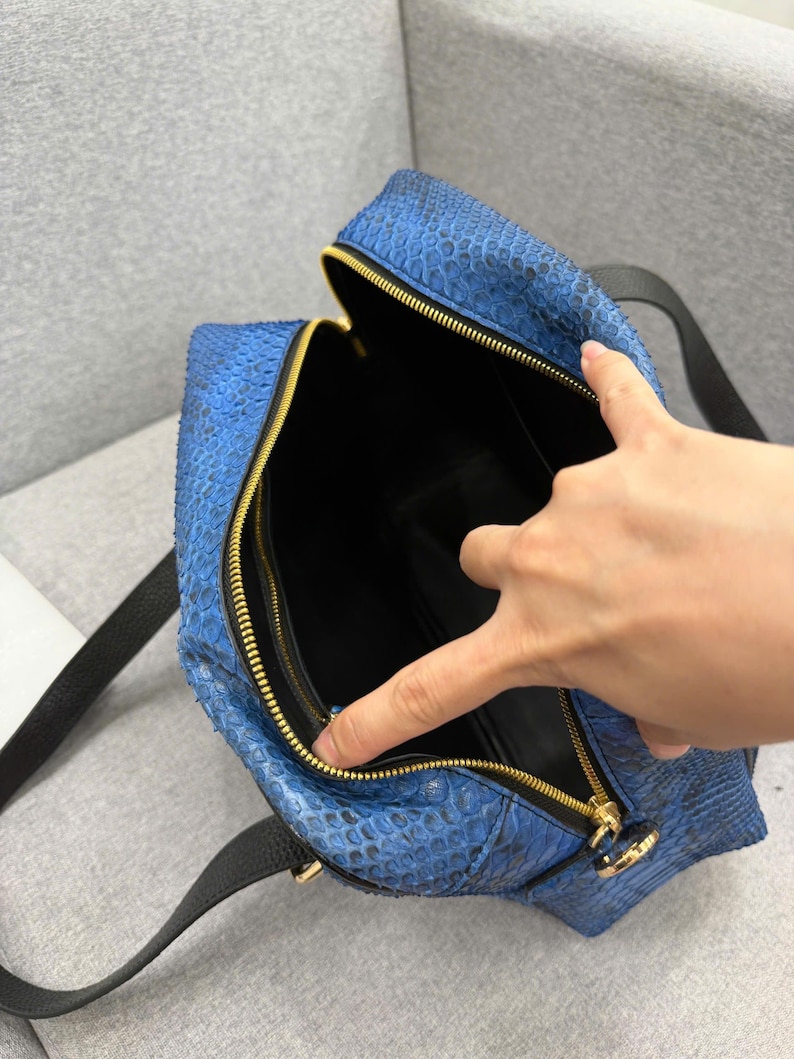 May include: A blue snakeskin-textured handbag featuring a gold-toned zipper closure and black leather straps. The interior of the bag is lined with black material, and the top is open to reveal the main compartment and a zippered side pocket.