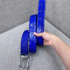 May include: A bright blue belt with a crocodile skin effect. The belt has a silver-coloured buckle. The item is displayed against a neutral background. The belt is a fashion accessory.