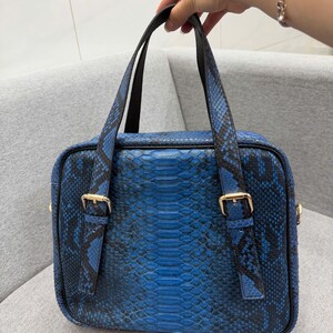 May include: A rectangular handbag featuring a vibrant blue and black snakeskin pattern. The bag includes two dark handles with gold-toned metal buckles on the front straps. The design has a structured shape with a zipper closure along the top edge.