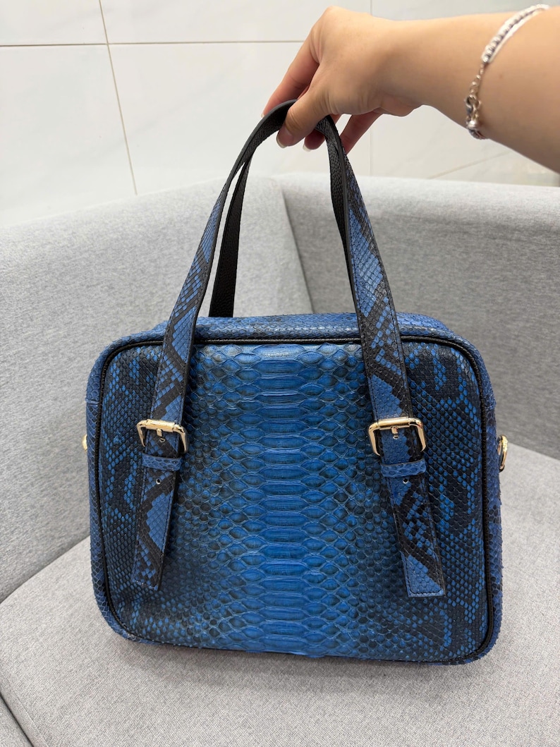 May include: A rectangular handbag featuring a vibrant blue and black snakeskin pattern. The bag includes two dark handles with gold-toned metal buckles on the front straps. The design has a structured shape with a zipper closure along the top edge.